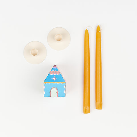 Honey colored taper candles, pair of off-white metal Hawkins New York Candle Holders, and State the Label box of matches in the shape of a house with colorful illustrations