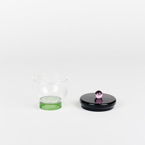 an Areaware Sophie Lou Jacobsen small glass salt bowl with a green footed base a black lid with a round lilac ball on top sitting beside it