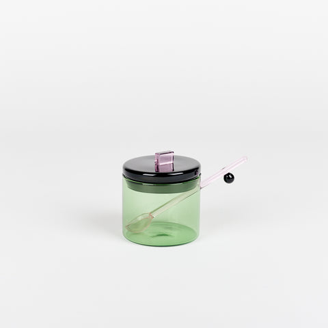 an Areaware Sophie Lou Jacobsen small green glass sugar bowl with a black lid with rectangular lilac handle on top and a small pink glass spoon inside