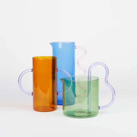 Three colorful glass vases positioned close together so that the colors - blue, green, and orange overlap. Each glass vase and pitcher has an interesting curved handle.
