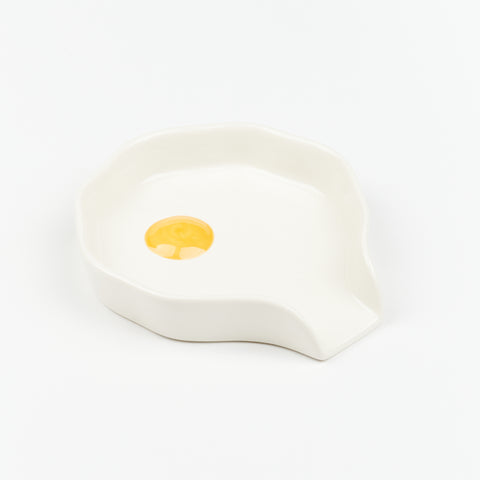 A circular white ceramic spoon rest with yellow circle in the middle made by Reva Preven