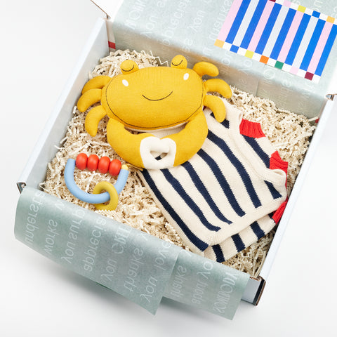 an open box featuring a baby gift that includes a plush smiling yellow crab toy, a organic knit sailor style baby romper onesie and a January Moon Teething Ring