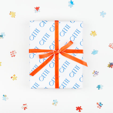 a box wrapped in Cette wrapping paper tied with an orange velvet ribbon surrounded by small colorful puzzle pieces