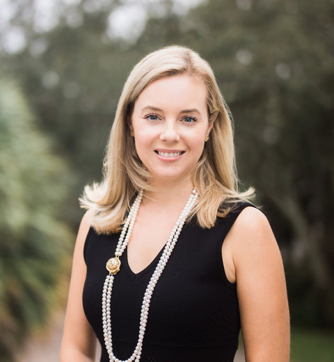 A young blond woman in a black dress wearing a long pearl necklace smiling and looking into the camera