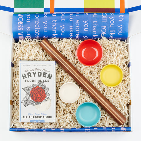 a gift box with blue tissue paper with words on it and in the gift box is a rolling pin, box of baking flour, and four measuring cups in different sizes and bright bold colors