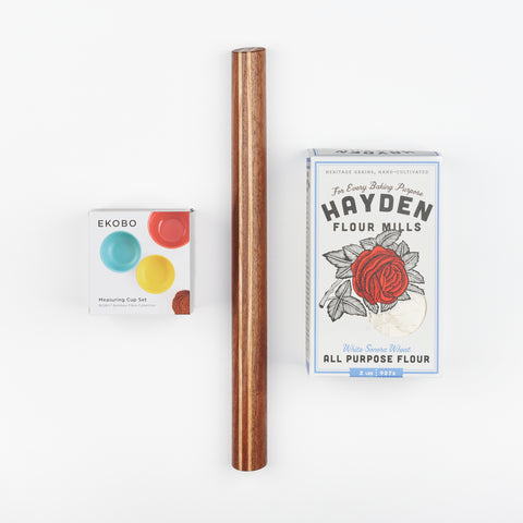 a box of measuring cups, a wooden rolling pin with two in-laid lighter wood lines and a box of Hayden Flour Mills All Purpose Flour