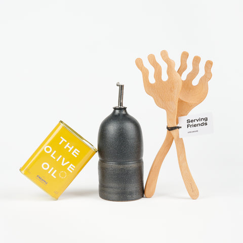 A Utility Objects black oil cruet between a yellow tin of olive oil and a pair of wooden serving spoons