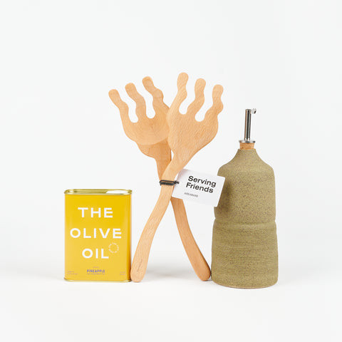 A pair of Areaware Serving Friends wooden tongs between a yellow tin of Pineapple collaborative Olive Oil and an olive colored ceramic cruet