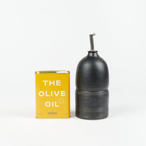 A black Utility Objects ceramic oil cruet with a metal pour spout standing upright next to a golden yellow tin of Pineapple Collaborative Olive Oil