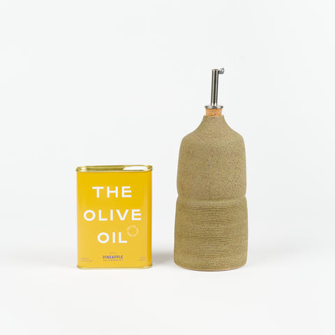 An olive colored Utility Objects ceramic oil cruet with a metal pour spout standing upright next to a golden yellow tin of Pineapple Collaborative Olive Oil