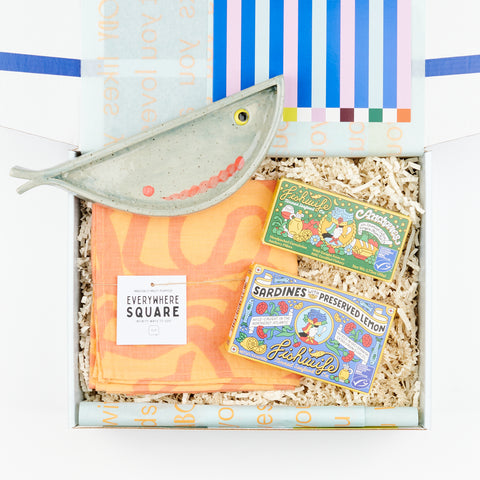 An open gift box showing a handmade ceramic plate in the shape of a fish, two boxes of Fishwife Tinned Fish, and a melon and pink Jenny Pennywood Everywhere home textile