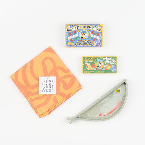 A Libby Hruska ceramic plate in the shape of a fish, two boxes of Fishwife Tinned Fish - Anchovies and Sardines, and a melon and pink Jenny Pennywood Everywhere home textile