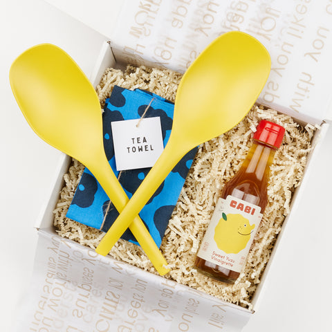 Two yellow salad tongs with a folded blue tea towel and a small bottle of vinaigrette all in a gift box