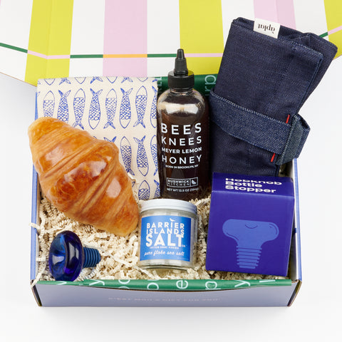 An open gift box with an assortment of items perfect for a new housewarming gift including an Areaware Hobknob bottle stopper, a Pampshade Croissant Bread Lamp and more