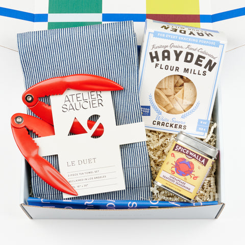 An open gift box showing two red lobster claw crackers, a box of white sonora crackers, a tin of Spicewalla Chesapeake Seafood Seasoning and two Hickory Stripe Tea towels by Atelier Saucier