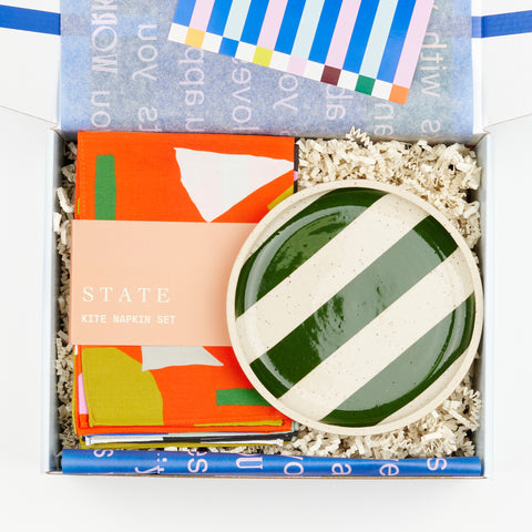 An open gift box with a handmade ceramic plate by Libby Hruska with dark green stripes and a set of State the Label Kite napkins