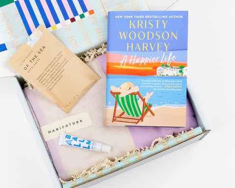 a color gift box showing a novel by Kristy Woodson Harvey, a purple Turkish towel, a packet of bath salts, and Zizia All Balm moisturizer