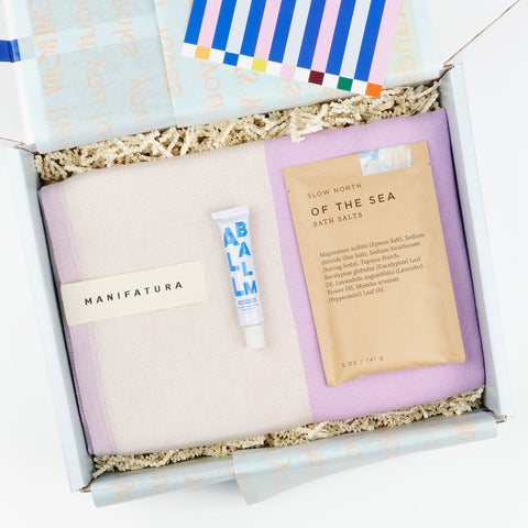 an open gift box featuring a gift set that includes a purple bath towel, a tube of all balm moisturizer and a packet of bath salts