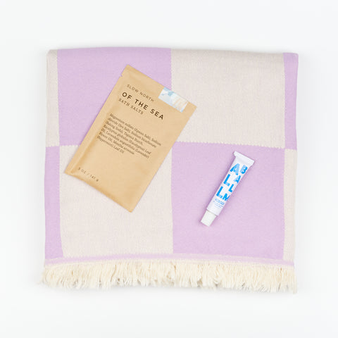 A brown packet of Slow North Of The Sea Bath Salts and a tube of Zizia All Balm moisturizer sitting on top of a purple Manifatura bath towel with a checkerboard pattern