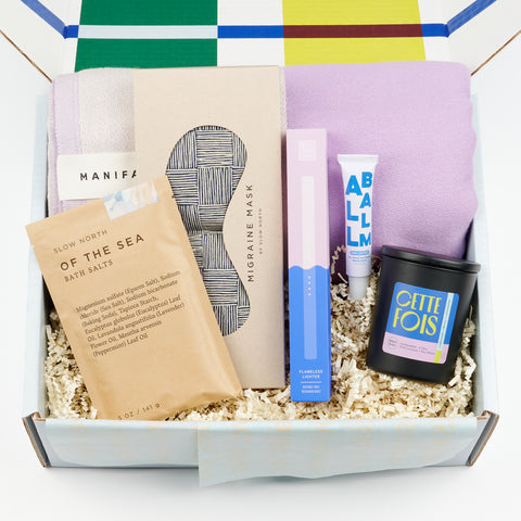 An open gift box for well-being including bath salts, a scented candle, a lavender bath towel, moisturizing balm, an eye mask, and a rechargeable flameless lighter