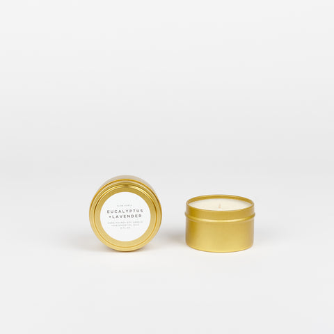 A Slow North Travel tin candle (Eucalyptus and Lavender scent) in a small brass colored tin with another candle in a tin beside it both are on a white background
