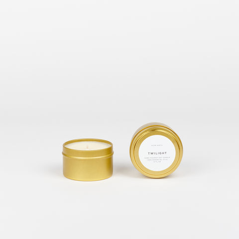 A Slow North Travel tin candle (Twilight scent) in a small brass colored tin with another candle in a tin beside it both are on a white background