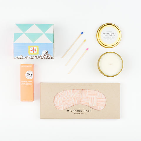 A Slow North Tin Candle, three matches, a State the Label box of House Matches in Vashon, a box of Lemon Laine Botanical Bounce Face Oil, and a Migraine eye mask with a pink crosshatch pattern