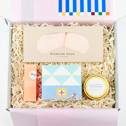 An open gift box featuring Lemon Laine Face Oil, an migraine mask, a house-shaped box of matches and a tin candle