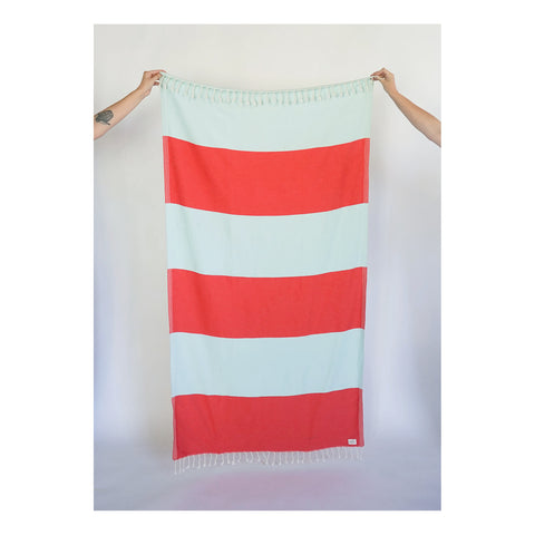 two arms holding up a bath towel with tassels and a red and light blue alternating large stripe pattern