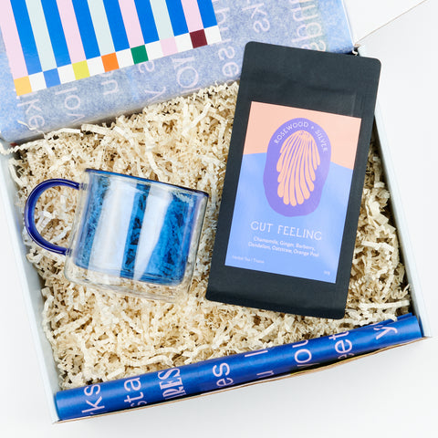 An open gift box showing a blue borosillicate double wall mug and a black bag of loose leaf tea called Gut Feeling