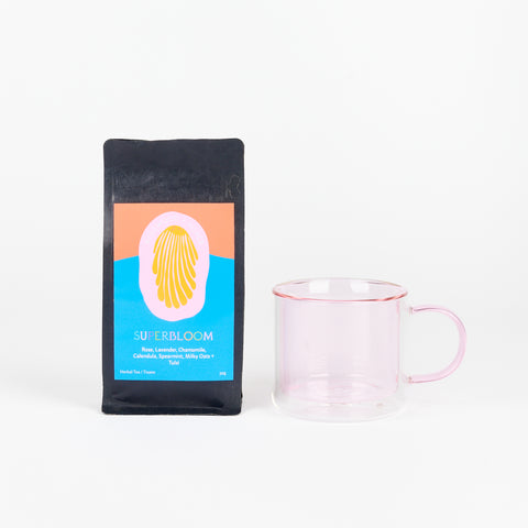 A Poketo Double Wall Mug in Pink next to a black bag of Rosewood + Silver Tea called Superbloom with a blue and orange label