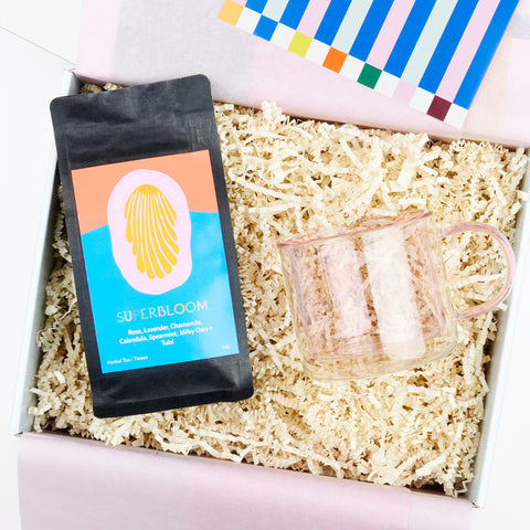 An open gift box with pink tissue paper showing a gift set that includes a black bag of loose leaf tea and a pink borosillicate glass double wall mug