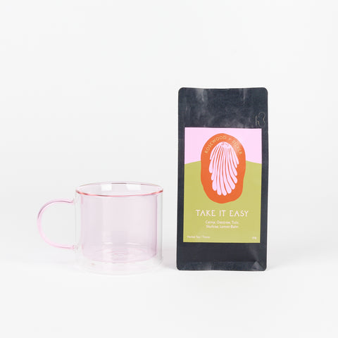 A Poketo Double Wall Mug in Pink next to a black bag of Rosewood + Silver Tea called Take It Easy with a pink and ochre label