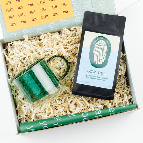 An open gift box featuring a gift set which includes a teal borosillicate glass mug and a bag of loose leaf tea