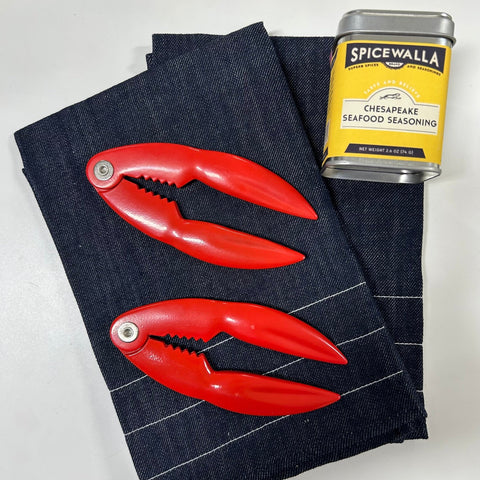 Two bright red lobster crackers resting on a park of dark denim tea towels, with a Spicewalla Chesapeake Seafood Seasoning tin placed nearby on a white background.