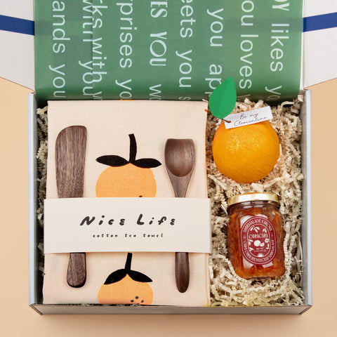 An open Jam On It (Holiday) gift box displaying artisanal marmalade, tea towel, and walnut spreaders, and a "be my clementine" surprise ball, all beautifully arranged