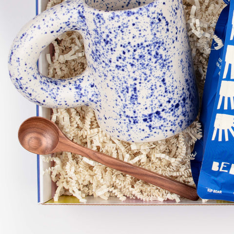 Close-up of the Morning Tide gift set, featuring walnut stirring spoon, speckled blue ceramic mug, and bag of small-batch coffee