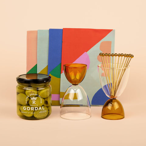 Front view of Olive Branch gift set showing two Mamo glasses, four multi-colored State the Label napkins, Regalis Gordal olives, and a set of A Bar Above cocktail picks arranged on a beige background.