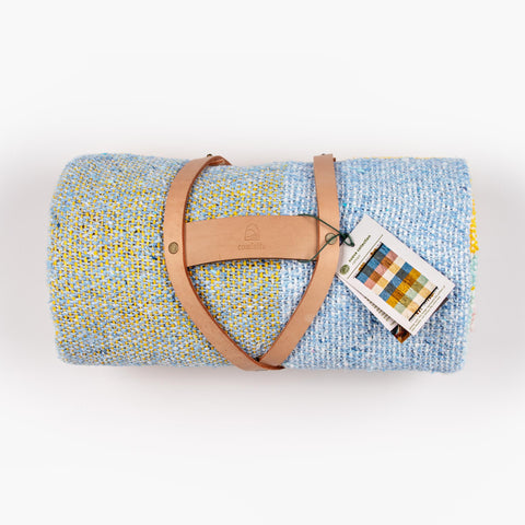 Handwoven Caminito blanket rolled with leather straps and carrying handle, included in the Kindled Comfort gift set. Shown from overhead on white background. 