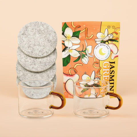 Steep Dreams gift set with Hudson Wilder glass mugs with amber handles, Woolly Made grey coasters, and Full Moon Tea Co. Jasmine Green tea on beige background.