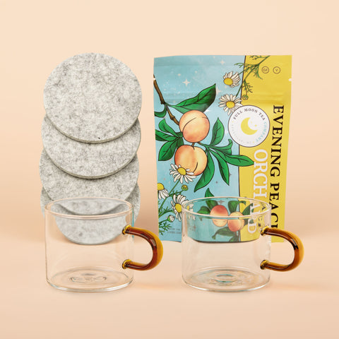 Steep Dreams gift set with Hudson Wilder glass mugs with amber handles, Woolly Made grey coasters, and Full Moon Tea Co. Orchard Peach herbal tea on beige background.