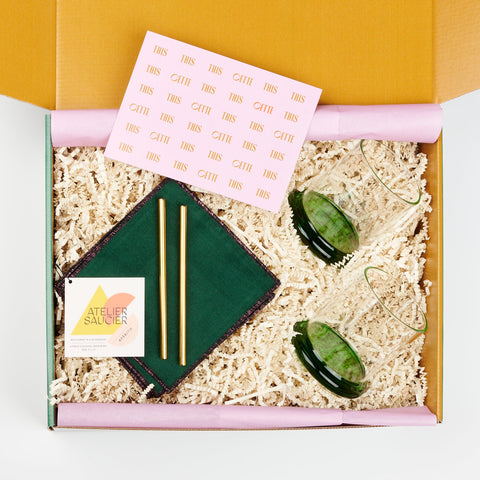 an open Cette gift box showing a pair of Sophie Lou Jacobsen fritter glasses with green bases, two metal cocktail straws and a set of four green velvet Atelier Saucier cocktail napkins