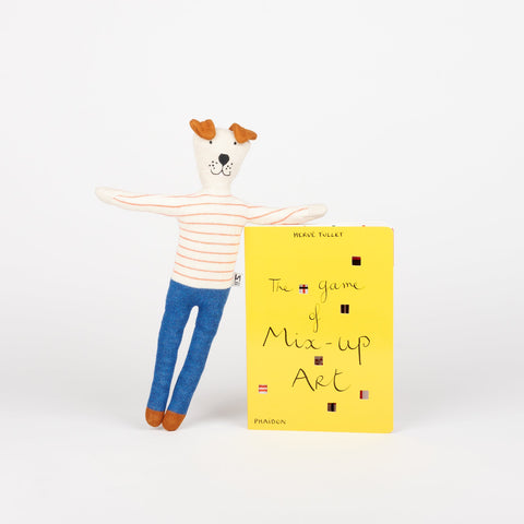 A Sophie Home stuffed animal toy dog in blue pants and red striped shirt standing and leaning on a copy of Phaidon Kids book The Game of Mix-Up Art a